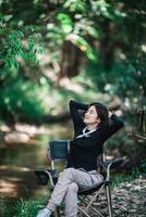 gelukkig jong vrouw zittend Aan stoel terwijl camping in Woud foto