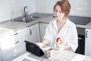 gelukkig mooi vrouw met kop van koffie of thee gebruik makend van laptop in quarantaine vergrendeling in de keuken in de wit shirt. foto