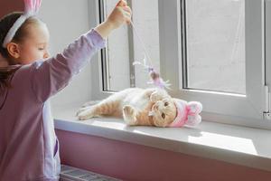 een schoolmeisje vervelend een hoofdband met konijn oren Toneelstukken met een gember huiselijk kat in een Pasen hoed Aan de vensterbank in de zon foto