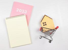 vlak lay-out van houten huis model- in boodschappen doen trolley met blanco bladzijde notitieboekje en roze dagboek of 2023 Aan wit achtergrond met kopiëren ruimte, huis aankoop plan concept. foto