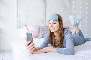 gelukkig jong vrouw ontspannende gebruik makend van en pratend met smartphone Aan bed Bij huis foto