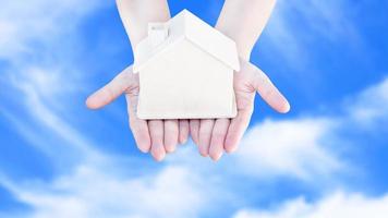 vrouw hand- Holding klein huis model- houten met wolk blauw lucht achtergrond. familie leven en bedrijf echt landgoed concept, eco huis foto