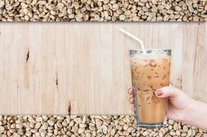 vrouw hand- Holding de glas bevroren koffie Aan houten en rauw koffie bonen achtergrond, bevroren latte koffie foto