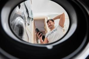 Mens met mobiel telefoon, visie van het wassen machine binnen. mannetje doet wasserij dagelijks routine. foto