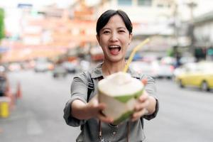 gelukkig jong Aziatisch vrouw rugzak reiziger drinken een kokosnoot sap Bij China stad- straat voedsel markt in Bangkok, Thailand. reiziger controle uit kant straten. foto