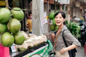 gelukkig jong Aziatisch vrouw rugzak reiziger staan in voorkant van kokosnoot sap winkel Bij China stad- straat voedsel markt in Bangkok, Thailand. reiziger controle uit kant straten. foto