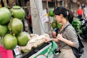 gelukkig jong Aziatisch vrouw rugzak reiziger staan in voorkant van kokosnoot sap winkel Bij China stad- straat voedsel markt in Bangkok, Thailand. reiziger controle uit kant straten. foto