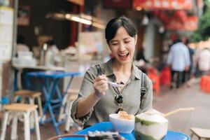 gelukkig jong Aziatisch vrouw rugzak reiziger genieten van straat voedsel Bij China stad- straat voedsel markt in Bangkok, Thailand. reiziger controle uit kant straten. foto
