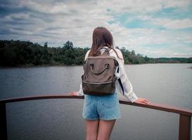 terug visie. jong Aziatisch eenzaam vrouw rugzak en staand Aan de brug aan het kijken de rivier- tussen bergen in zomer. ze was vervelend een wit shirt, denken over haar problemen en voelde heel verdrietig. foto