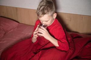 een gelukkig jongen is zittend in een rood bed onder een deken en drinken water van een glas foto