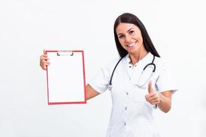 portret van een jong vrouw dokter in een laboratorium jas poses met een klembord in hand- tonen duimen omhoog. geïsoleerd Aan wit. foto