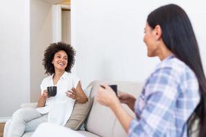 twee vrouw vrienden in sociaal afstand nemen zittend Aan bank. het beste vrienden hebben koffie samen terwijl gescheiden door sociaal afstand nemen Aan sofa Bij huis foto