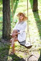 blond jong vrouw met slank naakt poten in wit jurk en rietje hoed zittend Aan een touw schommel foto