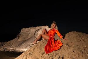 blond vrouw met slank naakt poten in oranje jurk zittend Aan de zand heuvel foto