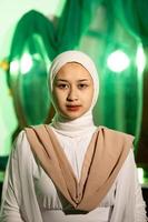 een moslim vrouw met een wit hoofddoek en wit kleren is zittend in de midden- van een groen kamer zonder bedenken foto