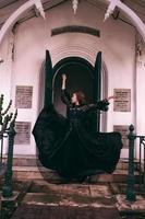 een elegant heks flicks haar allemaal zwart jurk in de lucht in voorkant van de begraafplaats poorten na de halloween festival begint foto