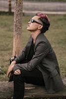 een Aziatisch Mens met zonnebril en een grijs blazer is genieten van de visie in de park foto