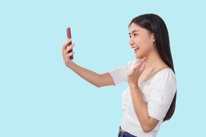 schattig Aziatisch tienerschattig Aziatisch tiener- meisje in wit overhemd staand gelukkig op zoek Bij smartphone terwijl video roeping met familie met blauw achtergrond. foto