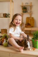 schattig weinig meisje in de houten keuken van de huis feeds de konijn vers gras foto