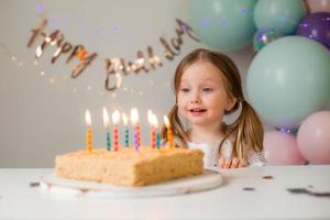 schattig weinig meisje slagen uit kaarsen Aan een verjaardag taart Bij huis tegen een backdrop van ballonnen. kind verjaardag foto