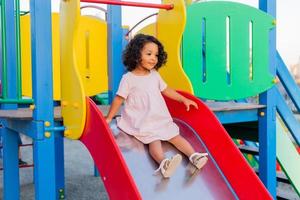 donker baby met gekruld haar- in een pale roze jurk Toneelstukken Aan een straat speelplaats en ritten naar beneden de heuvel. de concept van een gezond levensstijl. gelukkig jeugd. hoog kwaliteit foto