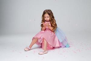 mooi weinig meisje in een chique roze jurk poses zittend Aan een grijs achtergrond en gooit confetti. mode en stijl. ruimte voor tekst. foto in de studio. hoog kwaliteit foto