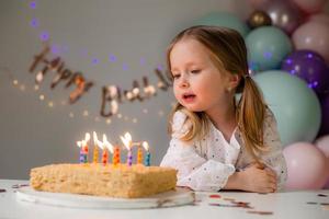 schattig weinig meisje slagen uit kaarsen Aan een verjaardag taart Bij huis tegen een backdrop van ballonnen. kind verjaardag foto