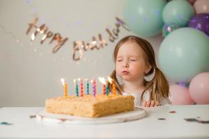 schattig weinig meisje slagen uit kaarsen Aan een verjaardag taart Bij huis tegen een backdrop van ballonnen. kind verjaardag foto