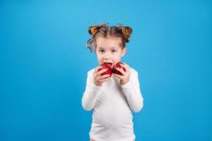 schattig weinig meisje met krullen in een gestreept jurk is Holding een groot rood appel in haar handen. blauw achtergrond. nuttig producten voor kinderen. gezond tussendoortje. ruimte voor tekst. hoog kwaliteit foto