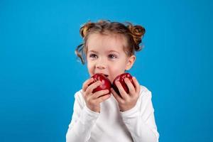 schattig weinig meisje met krullen in een gestreept jurk is Holding een groot rood appel in haar handen. blauw achtergrond. nuttig producten voor kinderen. gezond tussendoortje. ruimte voor tekst. hoog kwaliteit foto