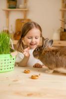 schattig weinig meisje in de houten keuken van de huis feeds de konijn vers gras foto