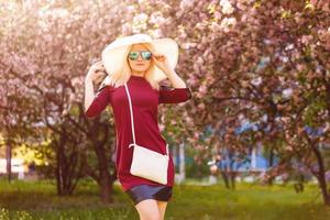 zomer mode portret van mooi blond vrouw poseren Aan verbazingwekkend bloeiend boom achtergrond .dragen zonnebril, gewoontjes hoed , modieus blauw zijde kimono en fluweel choker. elegant handtas. foto