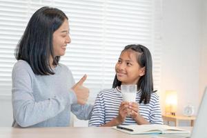 Aziatisch jong weinig meisje leren Bij huis. Doen huiswerk met soort moeder helpen, aanmoedigen voor examen. mam voorbij gaan aan Aan een glas van melk naar dochter. meisje gelukkig thuisschool. mam onderwijzen en advies onderwijs samen. foto