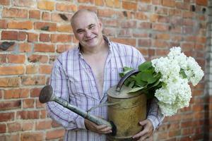 ouderen mannetje bloemist. een Mens houdt een gieter kan met een boeket van bloemen tegen een steen muur. passie voor de tuin. foto