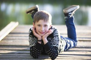 een knap jongen leugens Aan een houten brug en glimlacht. kind in natuur. foto
