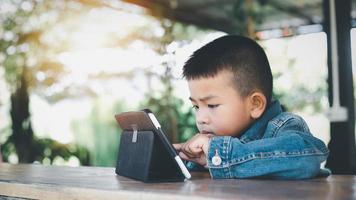 Aziatisch jongen op zoek Bij de tablet scherm aandachtig. overprikkeld kinderen concept. te veel scherm tijd. 5 jaren jongen aan het kijken videos terwijl TV is werkende.smartphone, internet verslaving concept. foto