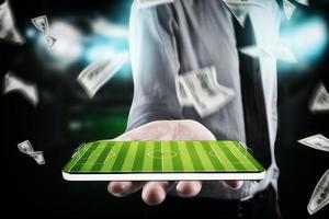 zakenman, met bankbiljetten in de omgeving van, houdt mobiele telefoon in zijn hand- met een Amerikaans voetbal veld- Aan de Scherm foto
