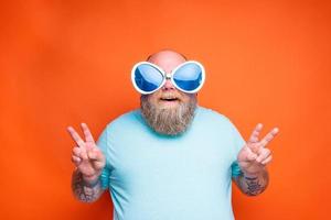 dik gelukkig Mens met baard, tatoeages en zonnebril maakt de teken van zege met zijn handen foto
