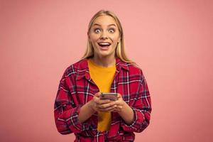blond schattig meisje types een bericht met haar smartphone. gelukkig en verrast uitdrukking gezicht. roze achtergrond foto