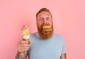 hongerig Mens met baard en tatoeages is onbeslist als naar eten een ijsje of een belegd broodje met Hamburger foto