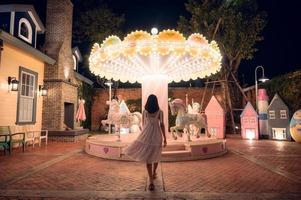 terug visie van Aziatisch vrouw in jurk wandelen naar verlichte carrousel Bij amusement park foto