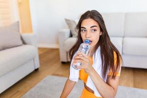 jong volwassen vrouw drinken water van plastic fles, zittend Aan geschiktheid mat en resting na opleiding Bij huis foto