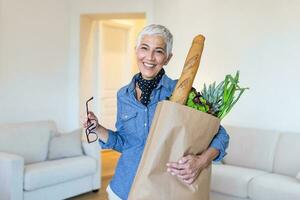 gezond positief gelukkig vrouw Holding een papier boodschappen doen zak vol van fruit en groenten. portret van mooi senior vrouw kruidenier boodschappen doen zak met groenten Bij huis foto