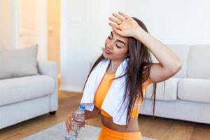 jong volwassen vrouw drinken water van plastic fles, zittend Aan geschiktheid mat en resting na opleiding Bij huis. mooi jong vrouw afvegen zweet met een handdoek, uitgeput na de dagelijks opleiding. foto