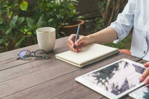 hand- vrouw schrijven notitieboekje Aan hout tafel met kop koffie en tablet. foto