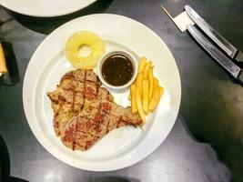 top visie t-bone varkensvlees steak met Frans Patat en pijnboom appel, peper saus Aan wit keramisch bord met steak messen Aan zwart leer tafel in de restaurant. foto