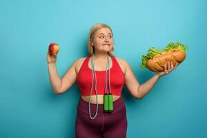 dik meisje is onbeslist naar eten een appel of een groot Sandwich. cyaan achtergrond foto