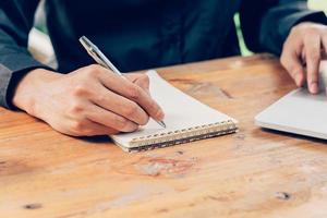 Mens hand- schrijven notitieboekje papier en gebruik makend van telefoon Aan hout tafel in koffie winkel met wijnoogst afgezwakt filter. foto