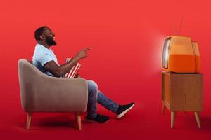 geamuseerd televisiekijker zittend in de fauteuil Bij huis terwijl aan het eten popcorns en aan het kijken een film Aan rood achtergrond foto