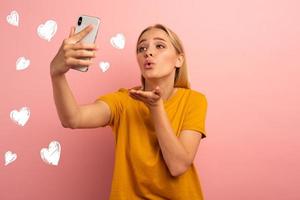 blond schattig meisje stuurt harten Aan haar smartphone. gelukkig en lief uitdrukking gezicht. roze achtergrond foto